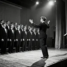 A vintage black and white photograph of the Moravian Teachers' Male Voice Choir performing on stage, led by a young conductor (Antonín Tučapský) with an energetic posture.